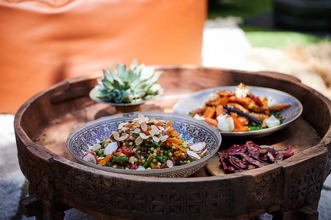 A colourful salad dish with couscous, sliced almonds, green beans, orange capsicum and sliced radish artfully displayed on a decorative wooden platter beside a second dish with orange vegetables and a succulent plant