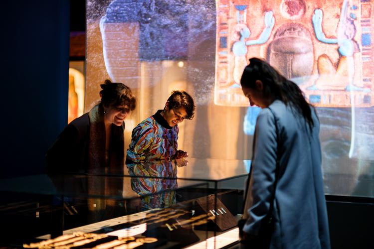 Three people wearing colourful clothing stare at a showcase containing small Egyptian amulets, with expressions of awe and interest while a large projection of an Egyptian pectoral creates a colourful background behind them