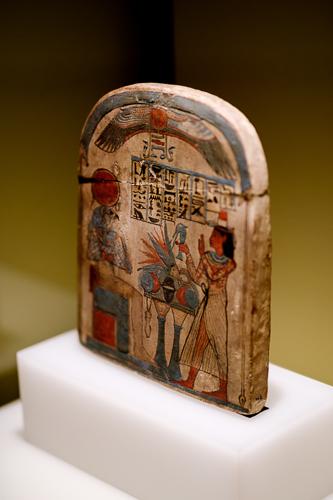 The Stela of Pamaaf, a small wooden block with a curved top edge, depicting a scene in which an Egyptian god with a hawk head sits at an offering table, receiving various offerings from an Egyptian man. 