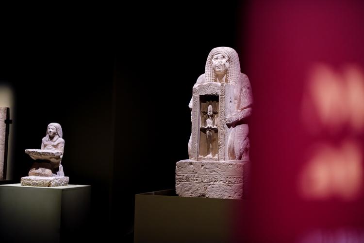 A large white statue on the right depicts Horemheb, in human form, wearing an intricate wig and knealing forwards while holding a rectangular carved box that contains a representation of a god in mummiform. On the left, a smaller statue is depicted kneeling and holding a small offering table.