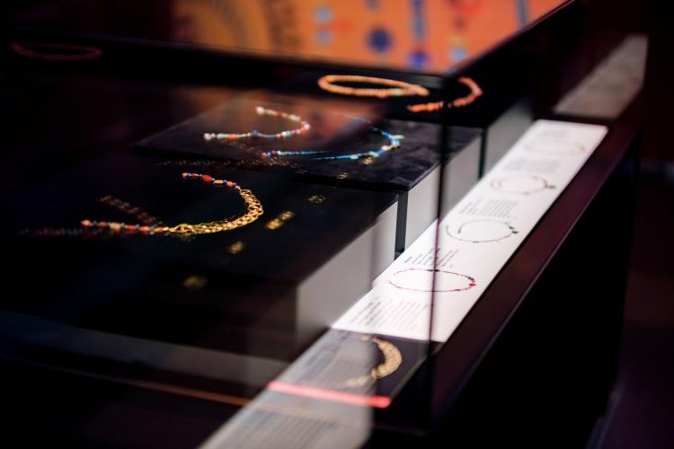 A mid shot of three black plynths each holding one to two beaded Egyptian necklaces. The necklaces range in colour, comprising beads of blue, red and orange and the necklace in the foreground appears to be adorned with tiny interwoven gold rings