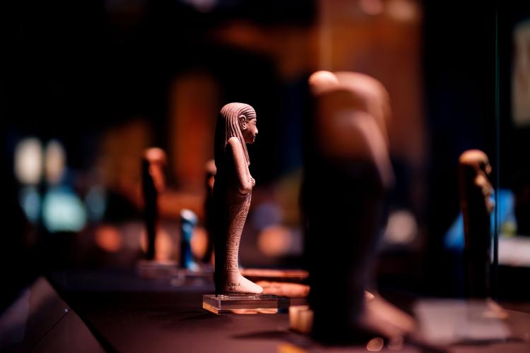A close up, side shot of a small brown Shabti, or Egyptian religious figure approximately fifteen centimetres tall in mummiform with its arms crossed across its chest and hieroglyphic script running up its body