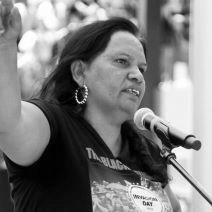An image of Megan Krakouer. An Aboriginal women speaking into a microphone. She has black hair and her hand raised, illustrating a speech.