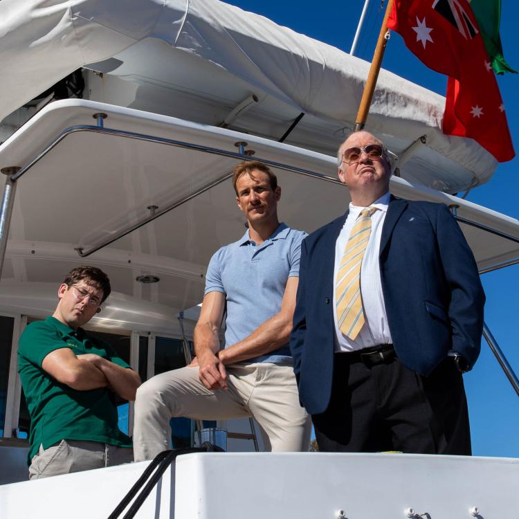 Three men, one dressed in a dark blue suit with a yellow tie and tinted glasses, and the other two in khaki slack pants and a blue and green polo shirt stand on a white boat and stare imposingly into the camera.