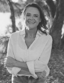 Image of a woman with shoulder length hair, wearing a white blouse, smiling standing next to a river. She wears a white shirt.
