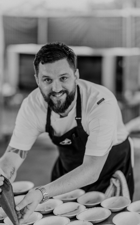 A black and white image of Executive Chef Stephen Watson who smiles into the camera as they pipe a pilling into empty pie shells
