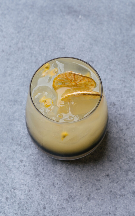 A yellow beverage in a short round glass garnished with dried citrus slices and passionfruit seeds