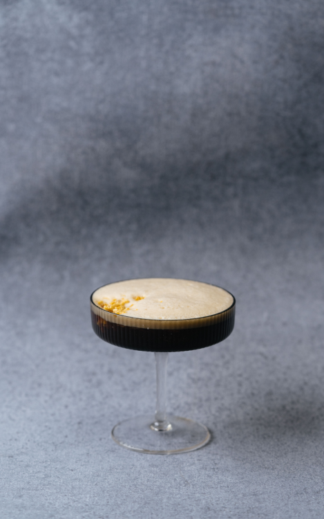A brown beverage with a foamy top and yellow garnish served in an aesthetic martini glass