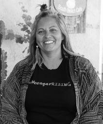 Image of an adult woman looking into the camera smiling. She has shoulder length hair, large feather earrings, and wears a shirt that says Noongar Rising.