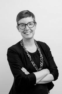 Image of a female identifying person with short hair, glasses, wearing a black blazer with her arms folded. She is smiling.