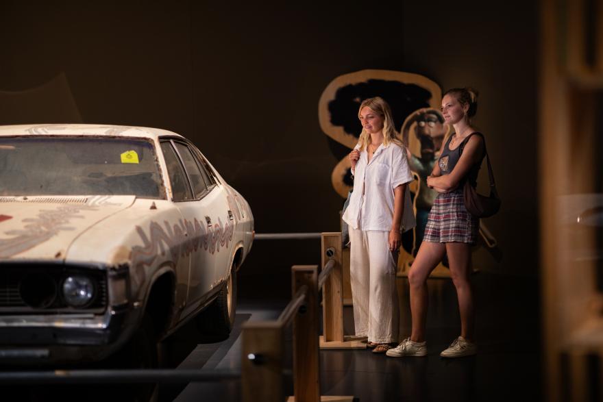 Two people in casual clothes stand at a barrier next to a white Fairlane car with curved artistic lines drawn along it by Aboriginal artists