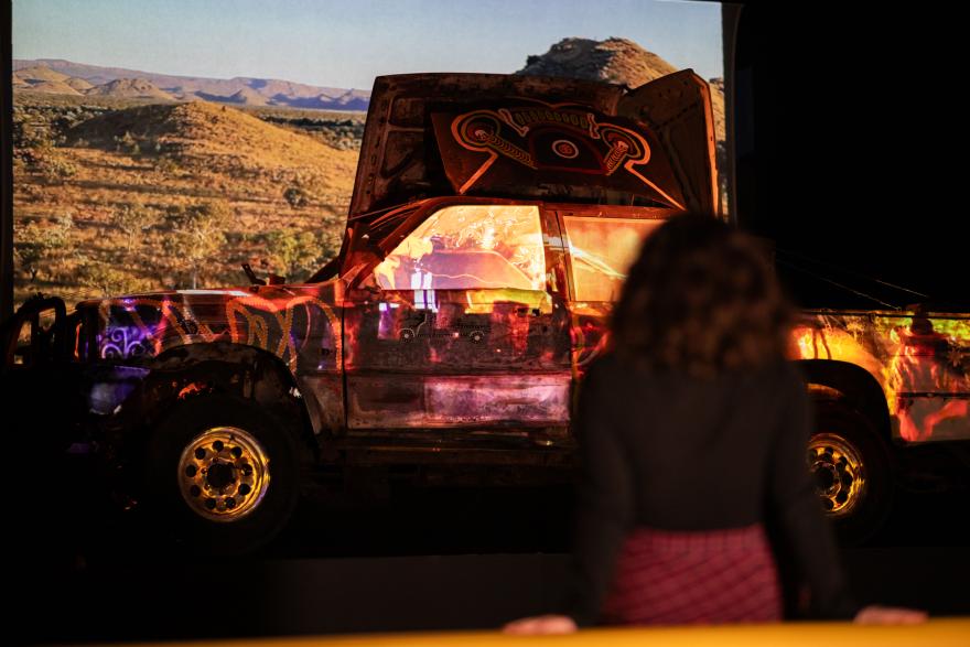 A person with curly hair and a black shirt sits on a couch slightly out of focus and watches a vividly-colourful projection and light show on an old car featuring images of country and bright paintings