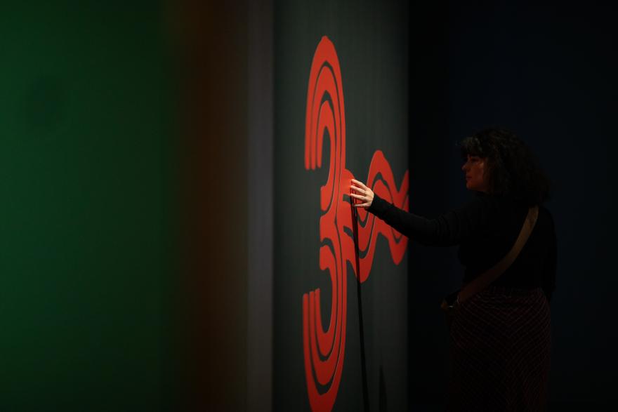 An artistic shot of a person mostly in silhouetted darkness reaching out to touch a graphic in the shape of a large numeric three made by three red squiggly lines.