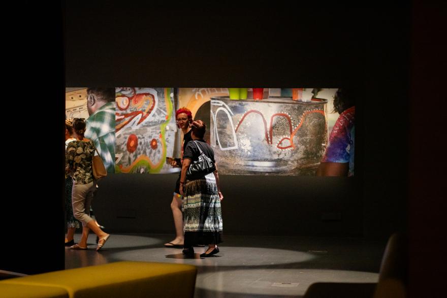 Several people converse with interested expressions as they walk past a long image of a red orange and white painting on a silver barrel