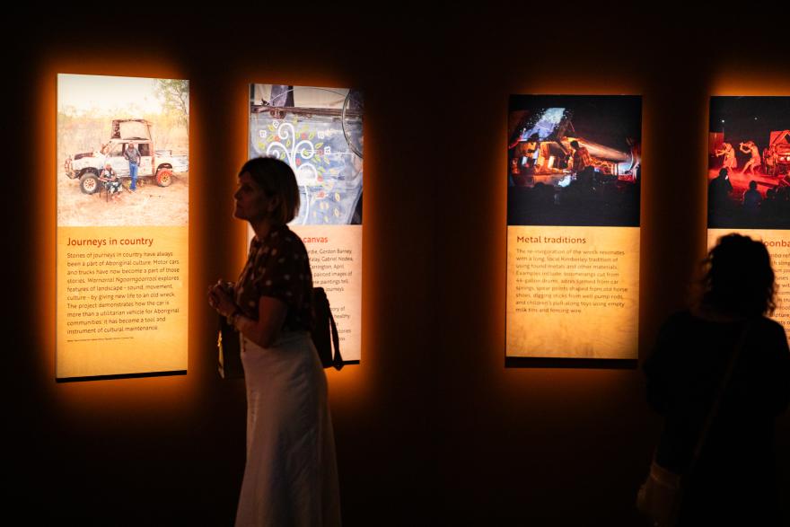 A person wearing a spotted shirt and long white skirt walks past several brightly lit graphic panels with images of artistic work in progress with red text on yellow writing beneath