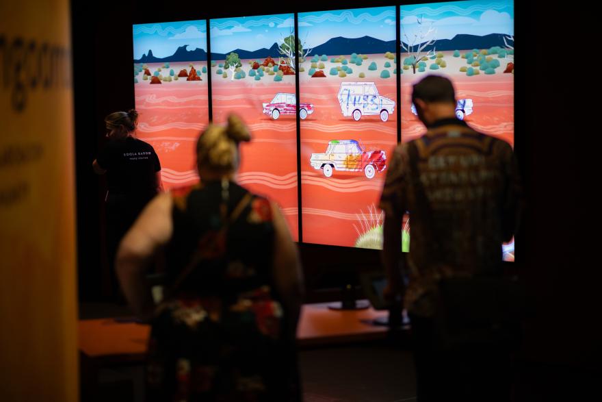 Several people stand in front of a large screen showing white trucks coloured with children's drawings racing down a red dirt cartoon road