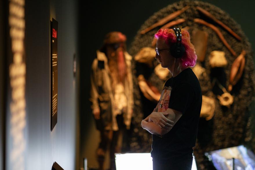 A person with orange and pink hair and a black t-shirt wears a pair of black headphones connected to an audio guide and stands with their arms crossed as though deep in though while reading from an exhibition sign