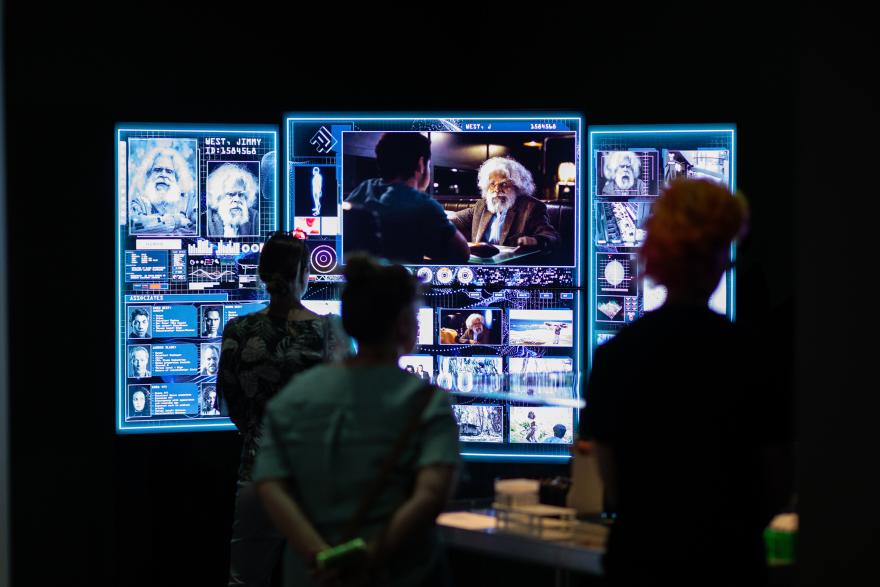 Several people stand with their backs to the camera looking at a bright screen styled like a surveillance spy information screen showing information about a character from the Cleverman series. It features surveillance shots, mug shots, written information, and mock medical information