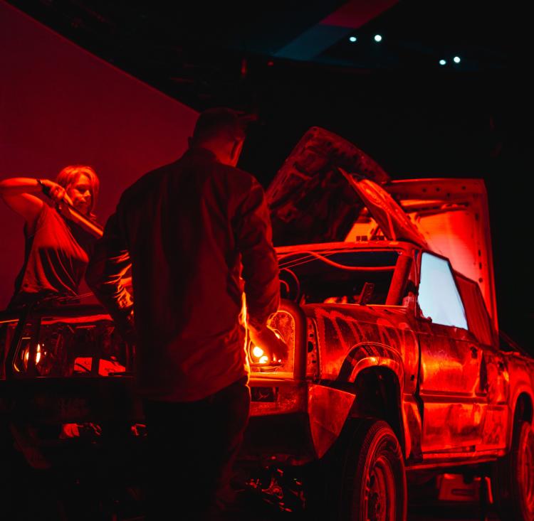 Vintage car illuminated by red lighting and intricate patterns, with two musicians playing beats on it 