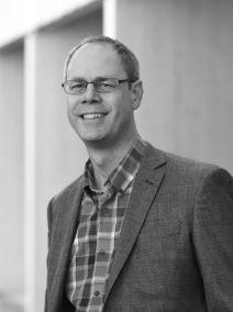 Image of a man wearing rectangular glasses and a suit jacket over a checkered shirt smiles into the camera with a warm expression.