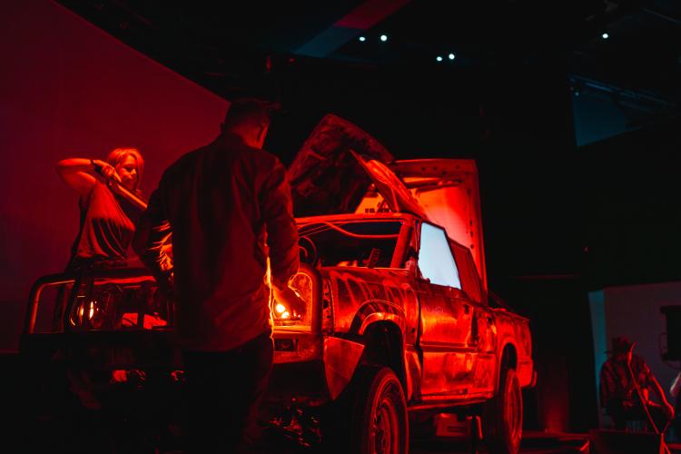An old car with Aboriginal paintings and artwork is bathed in a dramatic red light while several musicals stand around it with instrument tools in a performance