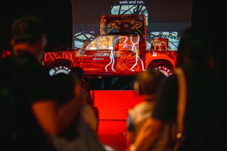 An old car lit with vividly colourful red white and yellow projections of drawings and videos is positioned on a pedestal as people gather around to watch it 