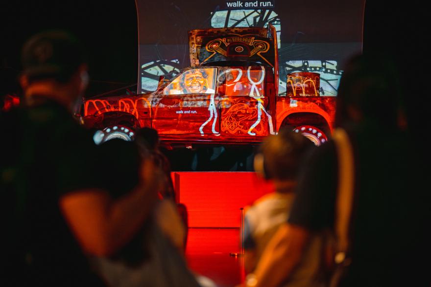 An old car lit with vividly colourful red white and yellow projections of drawings and videos is positioned on a pedestal as people gather around to watch it