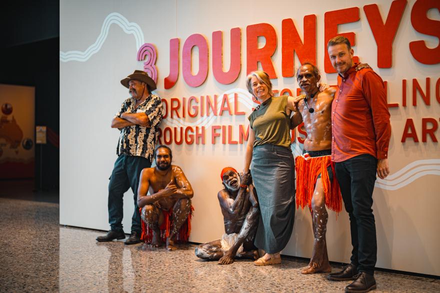 Several Aboriginal dances in orange skirts and white body paint pose with three people in casual jeans, button up shirts and skirts in front of the Three Journeys exhibition