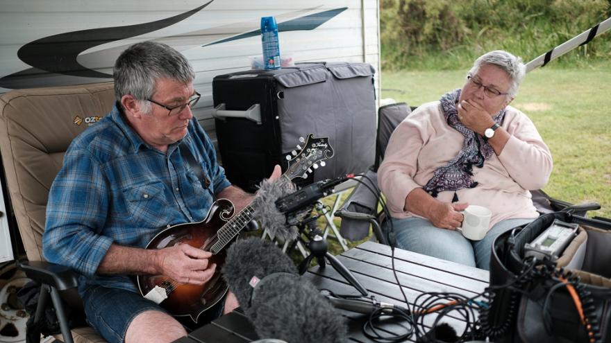 James and Dal Taylor sit on fold out camping chairs in front of a caravan in front of a table covered in recording equipment. James wears a blue checkered shirt and plays a small guitar-like instrument and Dal wears a pink shirt and purple scarf and watches James play.