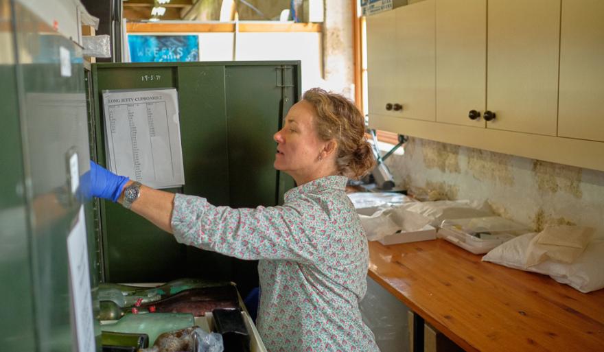 Corioli Souter wears a pink and green patterned button up shirt and blue object handing gloves and stares with concentration as they open a drawer of delicate objects at the Museum's collections storage facility.