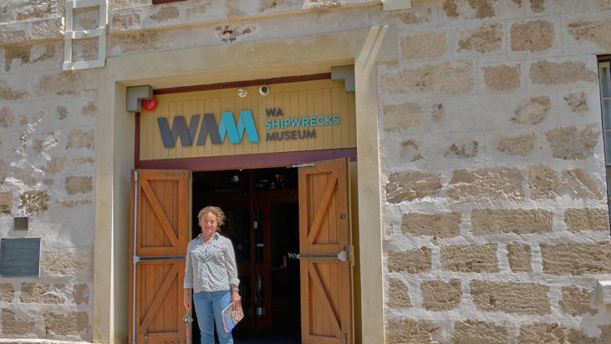 Corioli Souter wears a pink and green patterned button up shirt and blue jeans and stands in front of the wooden doors at the WA Shipwrecks Museum in Fremantle, holding a book in their hand