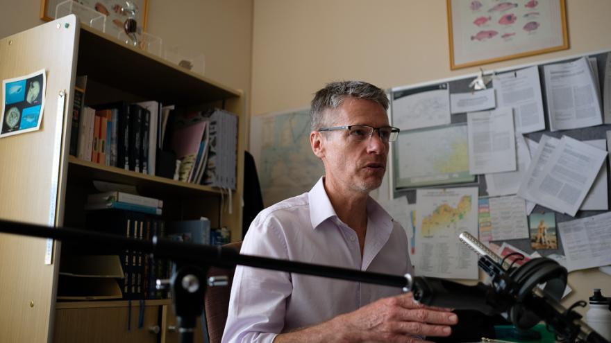 Glenn Moore wears a lilac button up shirt and thin framed glasses and gestures as though in mid conversation. They are sat in an office surrounded by books on bookshelves and paper pinned to a board