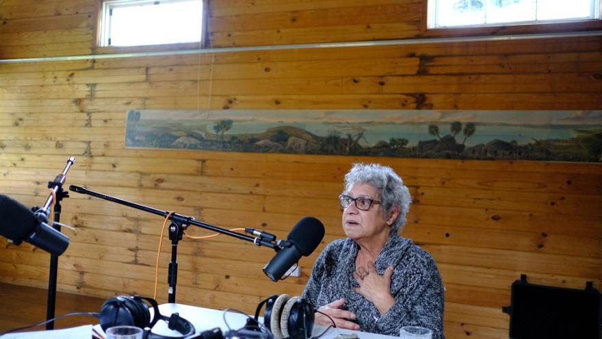 Vern Gillies wears a black and white patterned woollen jumper and sits at a table filled with podcasting equipment in a large wooden room. They hold their hand to their heart as though talking about something meaningful to them.