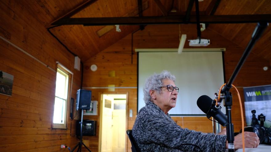 Vern Gillies wears a black and white patterned woollen jumper and sits at a table filled with podcasting equipment in a large wooden room. Their mouth is slightly open as though in mid conversation