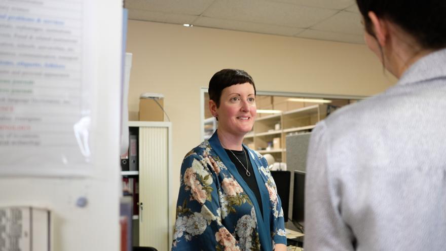Ali Clark wears a blue floral shawl over a black shirt and smiles as someone off camera as though in mid conversation. They stand in an office surrounded by computers and filing systems