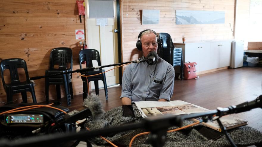 Malcolm trail wears a blue button up shirt and black headphones over their white hair as they sit at a table filled with podcast recording equipment in the middle of a wooden cabin with a fluffy microphone in front of their face