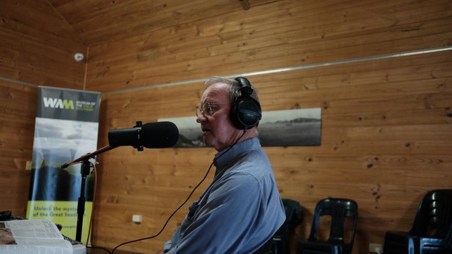 Malcolm trail wears a blue button up shirt and black headphones over their white hair as they sit at a table filled with podcast recording equipment in the middle of a wooden cabin with a fluffy microphone in front of their face