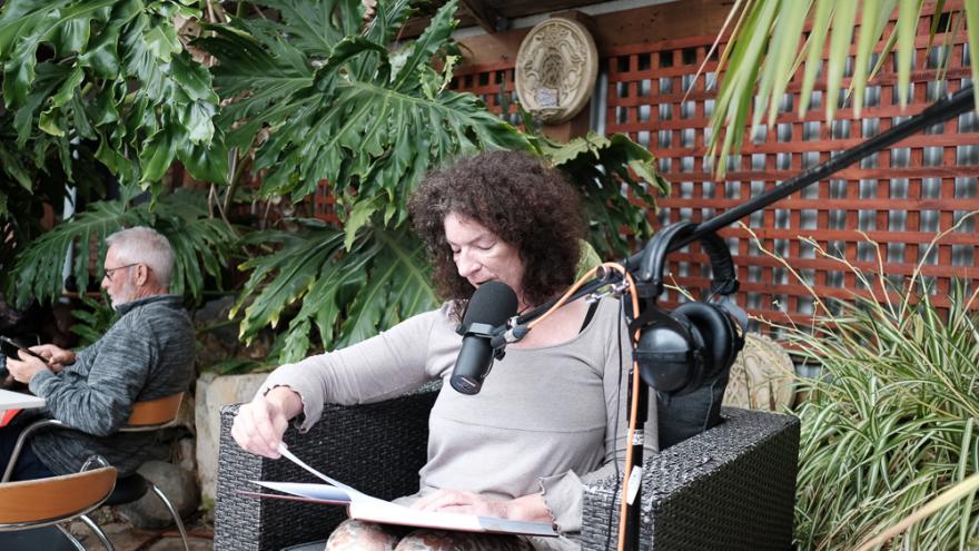 Sarah Drummond wears a grey long sleeve shirt with their curly brown hair falling around their shoulders and looks down at a book as they read a passage aloud with a black microphone near their mouth. They are seated in an outdoor cafe area surrounded by luscious green plants