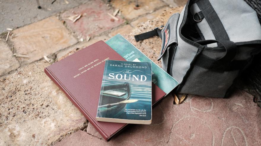 The book The Sound by Sarah Drummond with a blue cover featuring a wooden row boat is positioned on two larger research books next to a camera bag on a brick floor