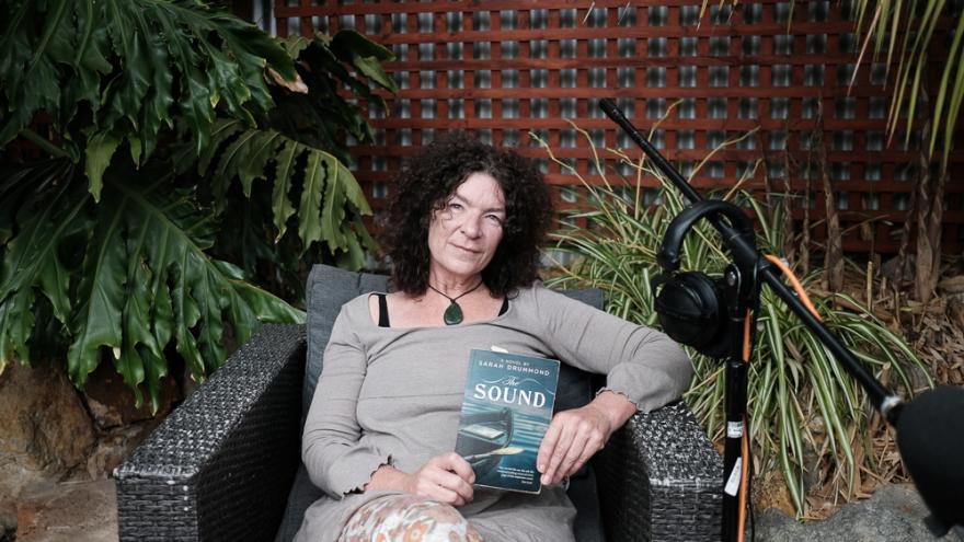 Sarah Drummond wears a grey long sleeve shirt and a large green pendant necklace with their curly brown hair falling around their shoulders and smiles at the camera as they hold a copy of their book The Sound up to the camera. They are seated in an outdoor cafe area surrounded by luscious green plants