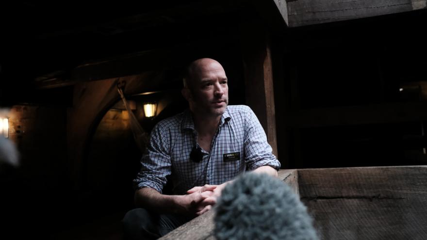 Scott Neil wears a white checkered shirt and learns against a wooden wall in the Brig Amity ship with their hands clasped as though discussing a serious topic.