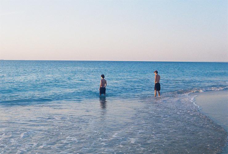 Two people in swimming shorts at a beach, walking into the ocean. The ocean is a deep blue and purple against a light blue and yellow dusk skyline