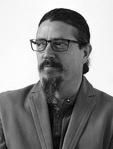 A black and white headshot image of Oron Catts who wears a blazer jacket over a button up shirt and black wireframed glasses. His chin-length beard and moustache is neatly combed and his hair is tied back in a bun, he looks off to the side of the camera