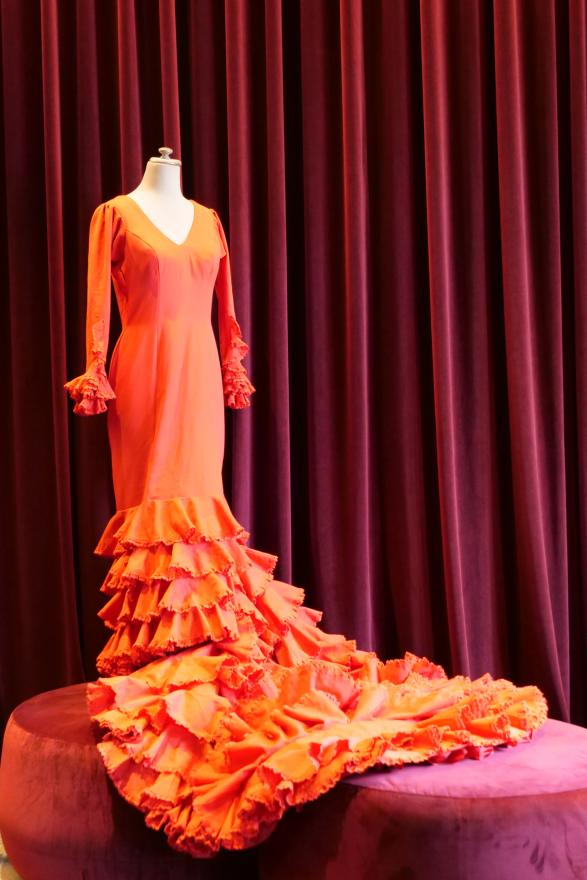 A red dress with a v-cut front and a long sleeves and skirts. The sleeves and skirt contains multiple rows of red ruffles. The costume is worn on a white mannequin in front of a red curtain.