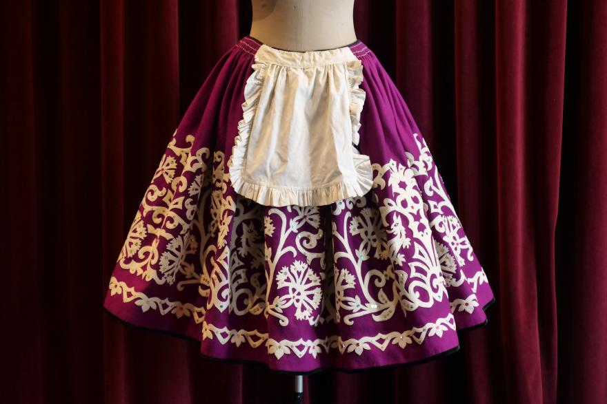 A purple skirt with a full shape and white detailing in a celtic-pattern style with a small frilly white bib. The skirt is worn on a white mannequin in front of a red curtain.