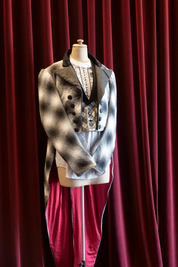 An evening jacket-style with a thick black collar and and black buttons with a black and white checked pattern and red velvet tails. The costume is worn on a white mannequin in front of a red curtain.
