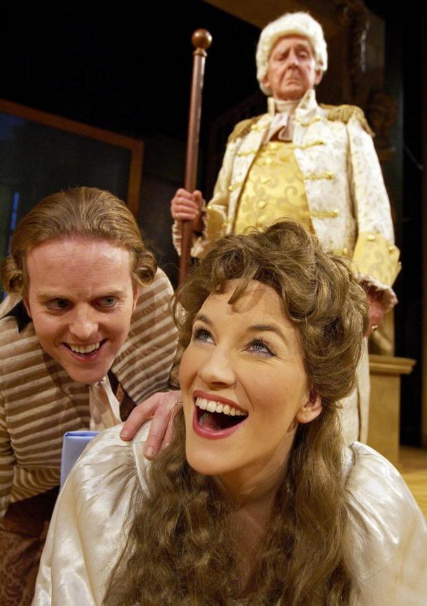 A person with curly brown hair and satin dress smiles up at something while another person with a curled wig kneels behind them with a hand on their shoulder. A third person in an ornate gold and white costume stands back from the couple and looks down at them