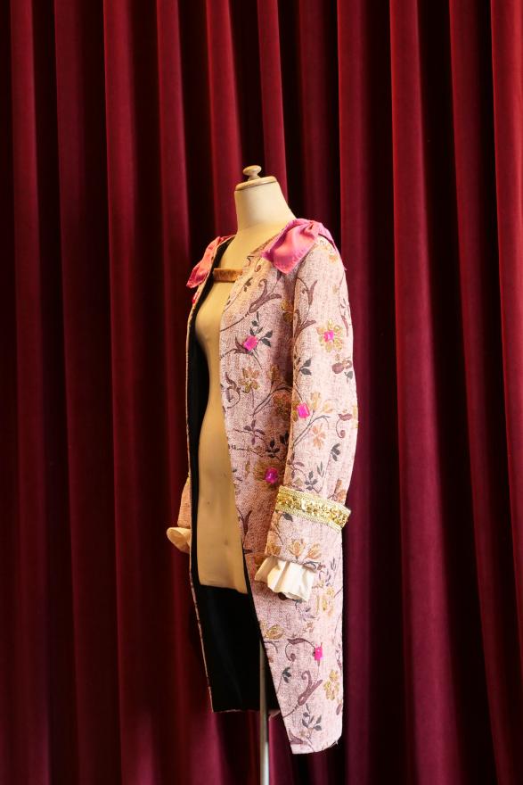 A light pink coloured regency-era jacket with a pink collar and gold detailing with hand-embroidered flowers. The costume is worn on a white mannequin in front of a red curtain.