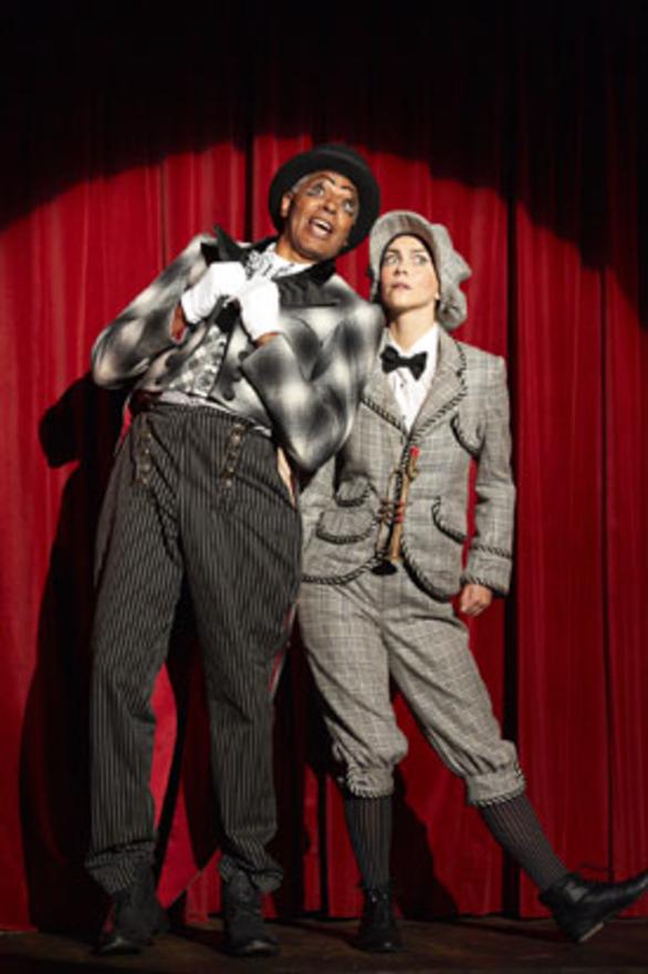 Two people in elaborate historic costumes in black, grey and white colours stand in front of a red curtain under a spotlight. The strike a pose as though talking to each other.