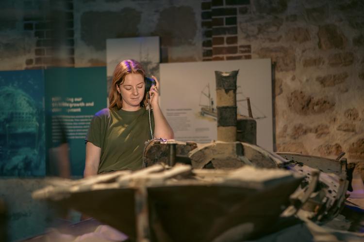 A person with long auburn hair wearing an olive green t-shirt holds a black audio puck to their ear and listens to a part of the exhibition. They stand behind a large model of a sunken ship and are surrounded by blue and white graphic panels and heritage brick work